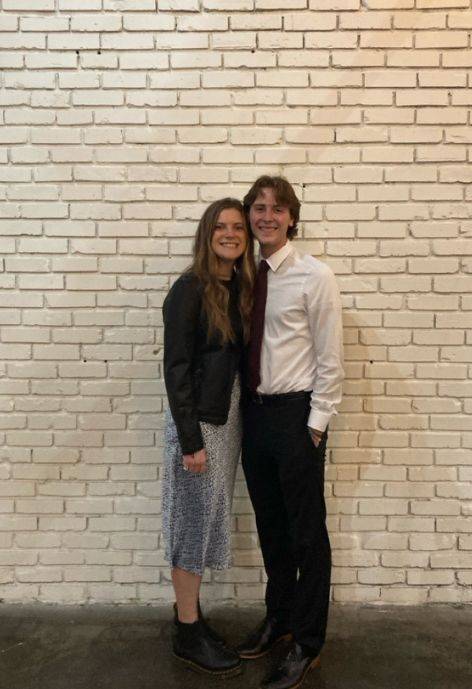 Daniel standing with a woman and smiling at the camera, there is a white brick wall in the background.