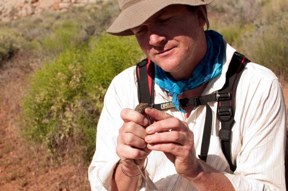 Man in a hat, looking down at his hands. He is holding a small lizard.
