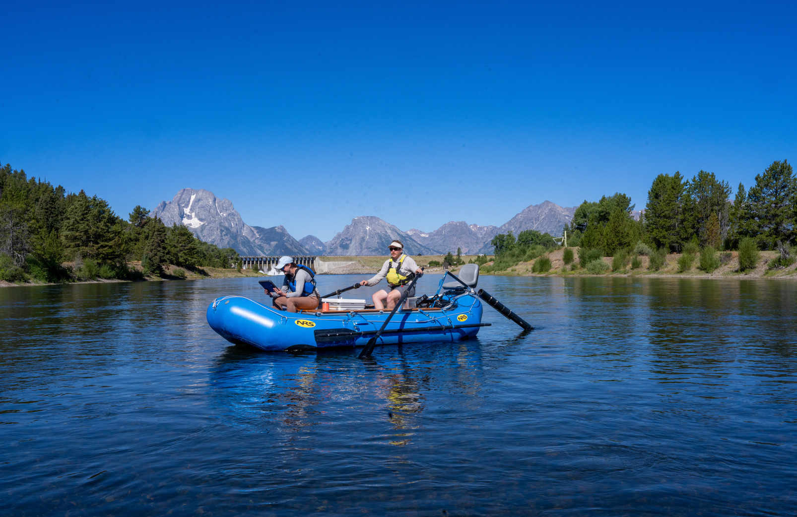 Two graduate students drive a boat on a blue lake, mountains in the background.