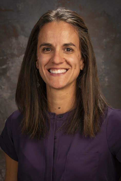 Nellie Bruns smiles while wearing a purple shirt and standing in front of a grey background.