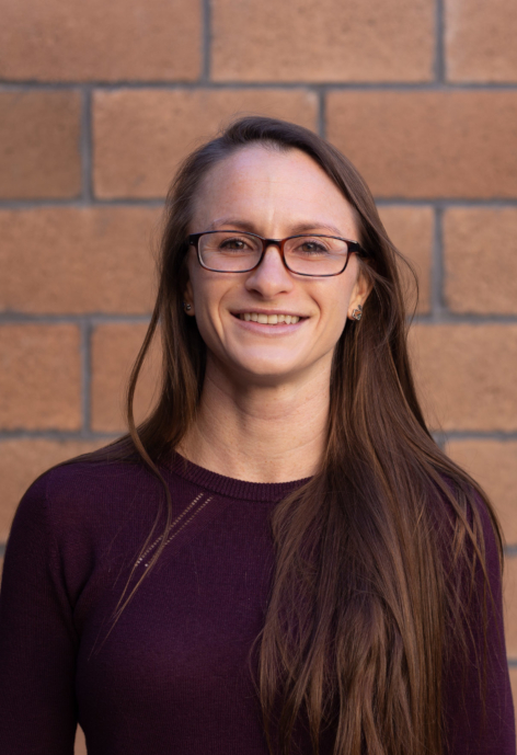 Jessie Williamson smiles in a maroon long-sleeve shirt in front of a brick wall.